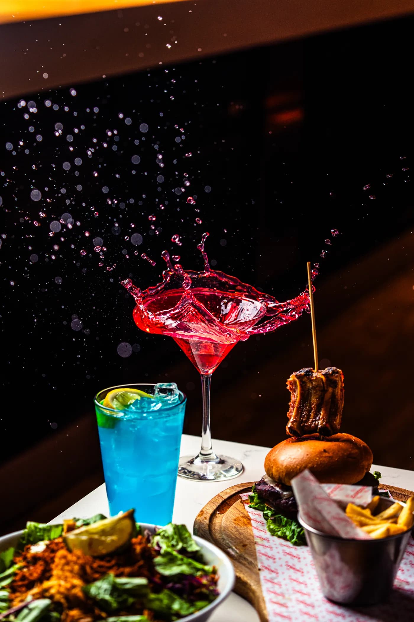 Coctel rojo con salpicadura junto a una bebida azul, una hamburguesa y una ensalada